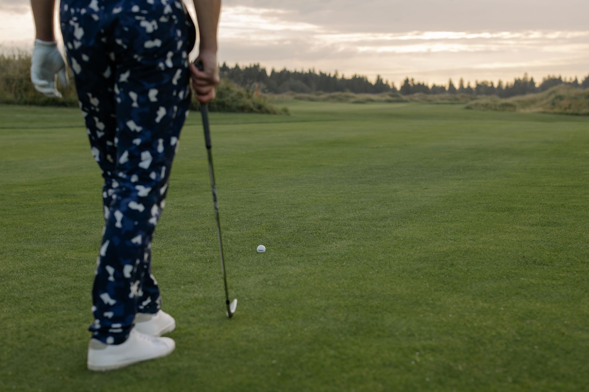 Golfer standing on a putting green, ready to putt at sunset, wearing patterned pants.