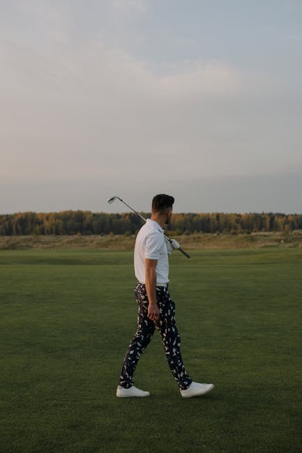 A stylish man walking on a serene golf course carrying a club over his shoulder.