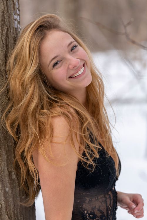 Free Young smiling female wearing black bodysuit looking at camera while standing near tree trunk in snowy nature on blurred background Stock Photo
