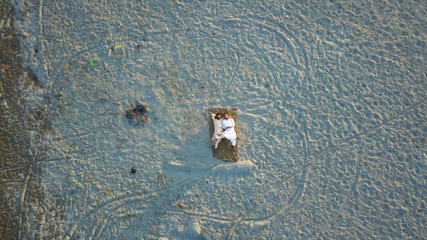 A couple resting on a blanket in an expansive sandy area viewed from above.