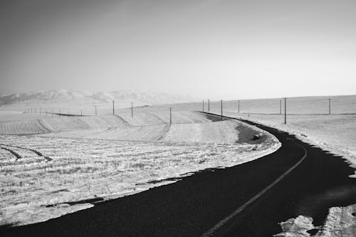 Black and white of empty asphalt roadway running through mountainous terrain with fields covered with snow