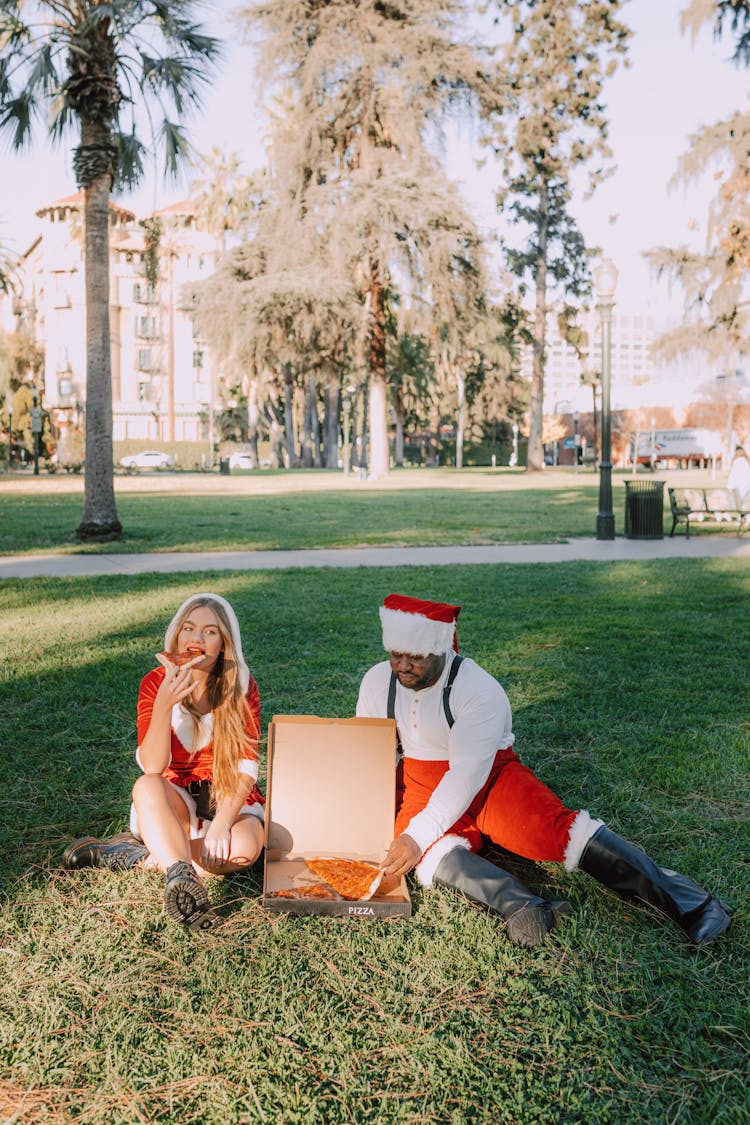 Man And Woman In Santa Costume Eating Pizza On The Grass