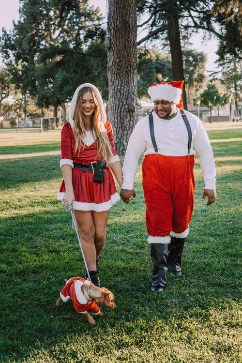 Free A joyful couple dressed in Santa costumes walking with a dog in a sunny park. Stock Photo