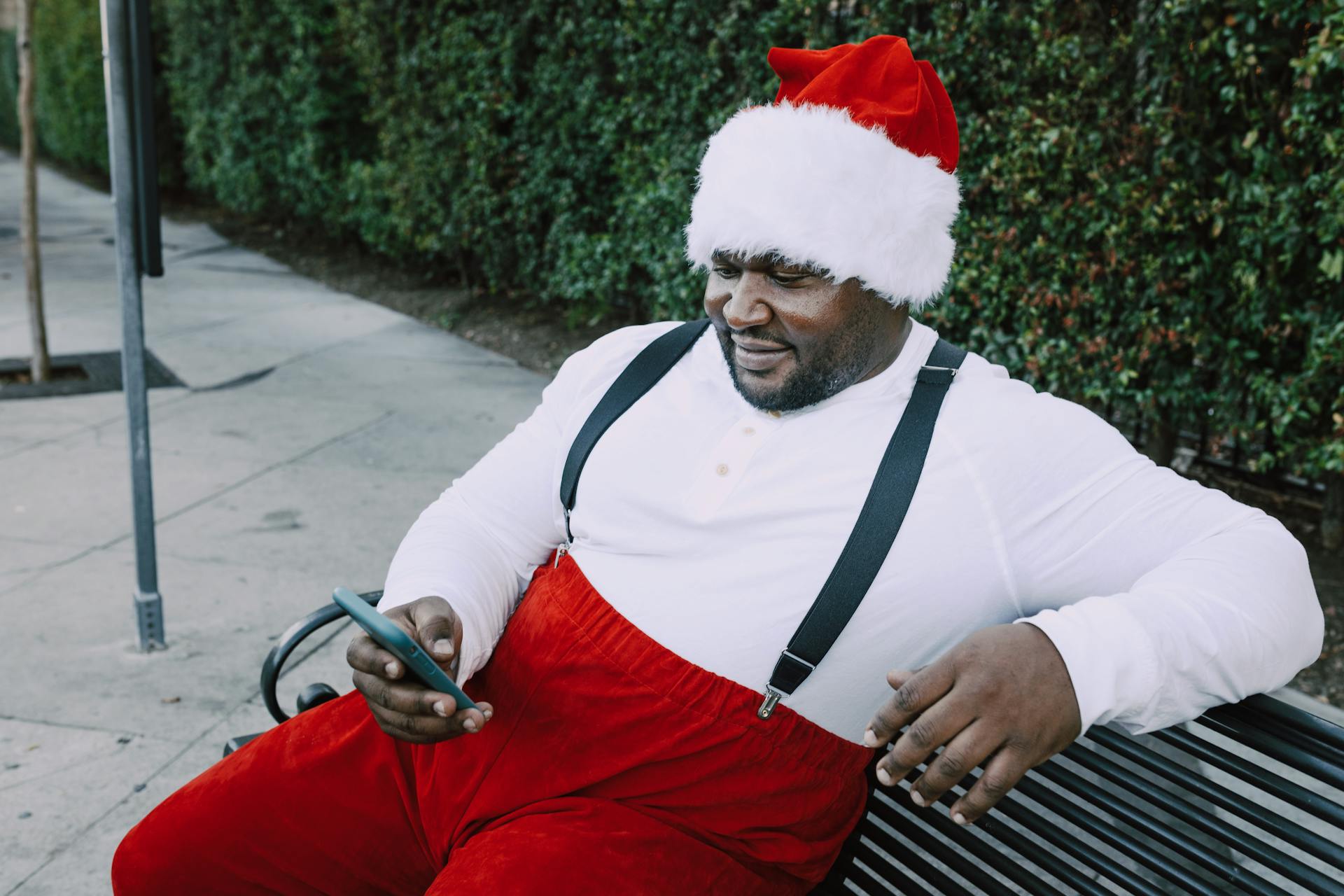 A man dressed as Santa Claus sitting on a bench using his phone outdoors, capturing a festive moment.