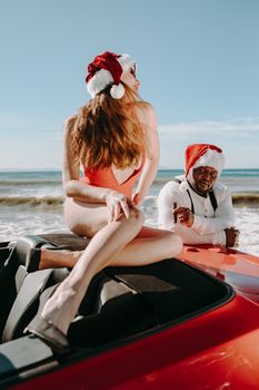 A man and woman in Santa hats enjoy a sunny day at the beach with a convertible car.