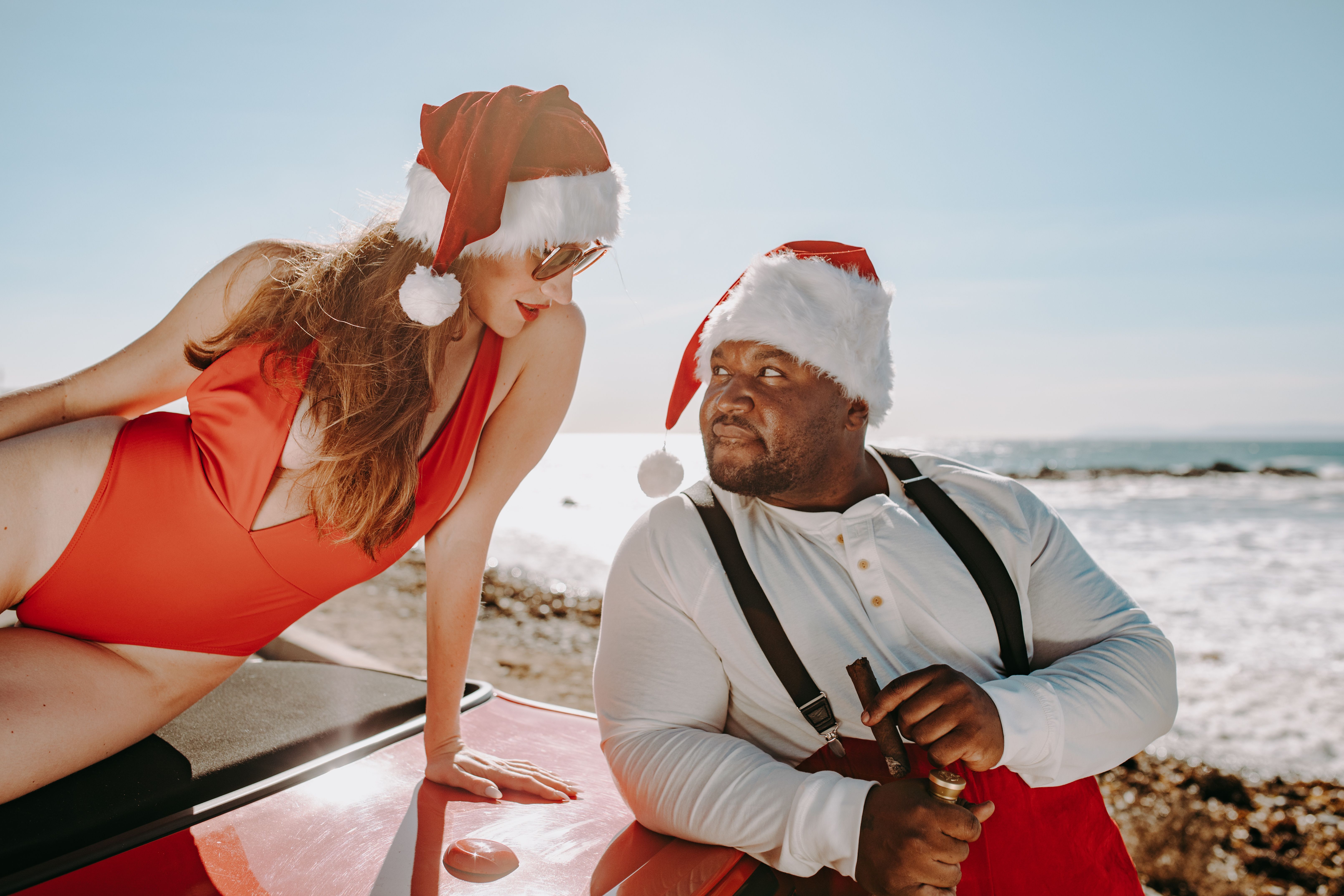 A couple celebrating Christmas at the beach in Santa hats and festive attire.