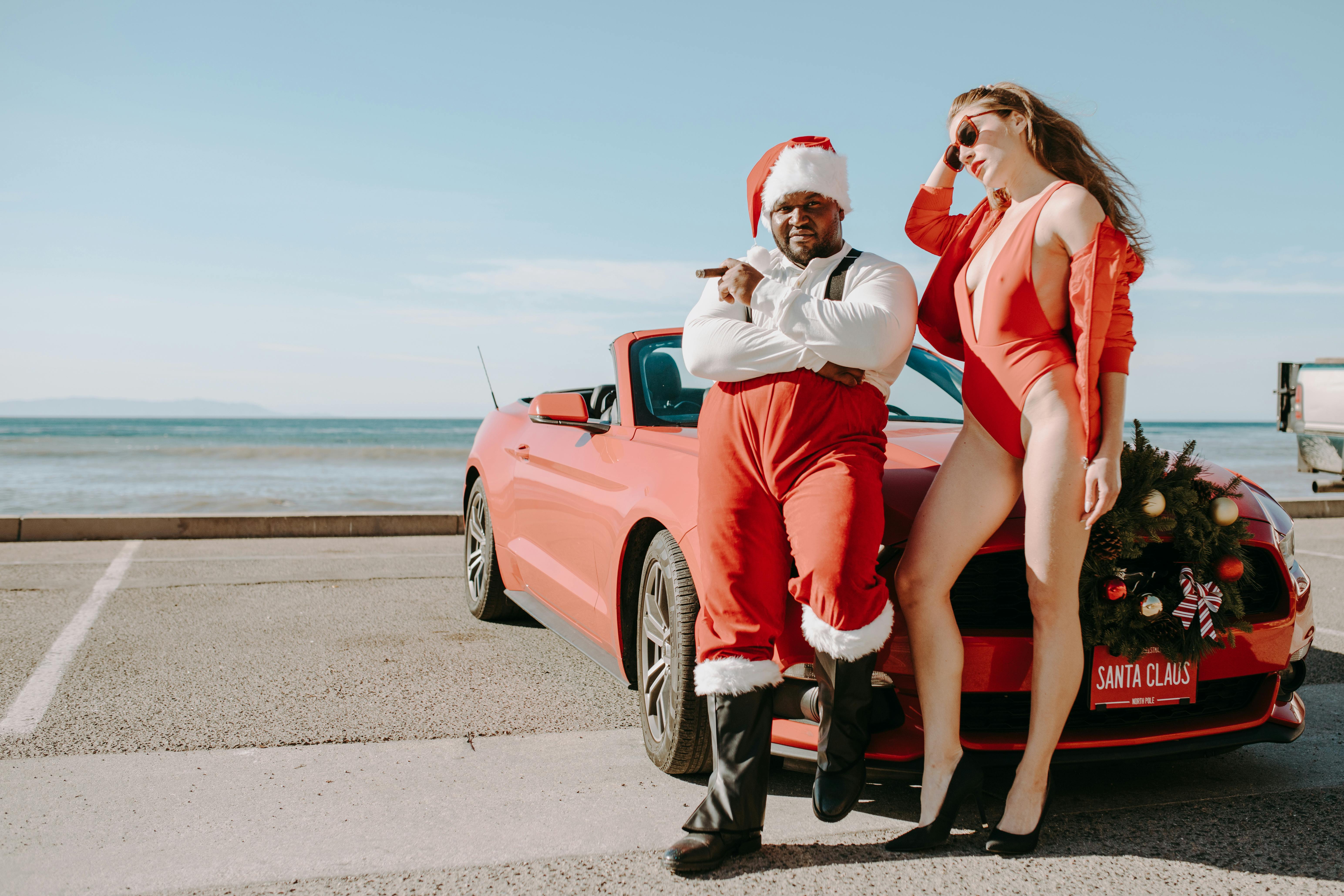 man in santa clothes while the woman in bikini standing in front of a car