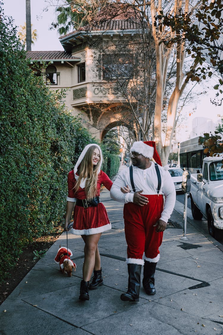 Man And Woman In Santa Outfit Walking On Sidewalk With Dog