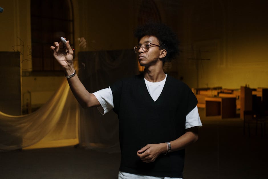 A young black man with afro hair writing on a transparent surface indoors under warm lighting.