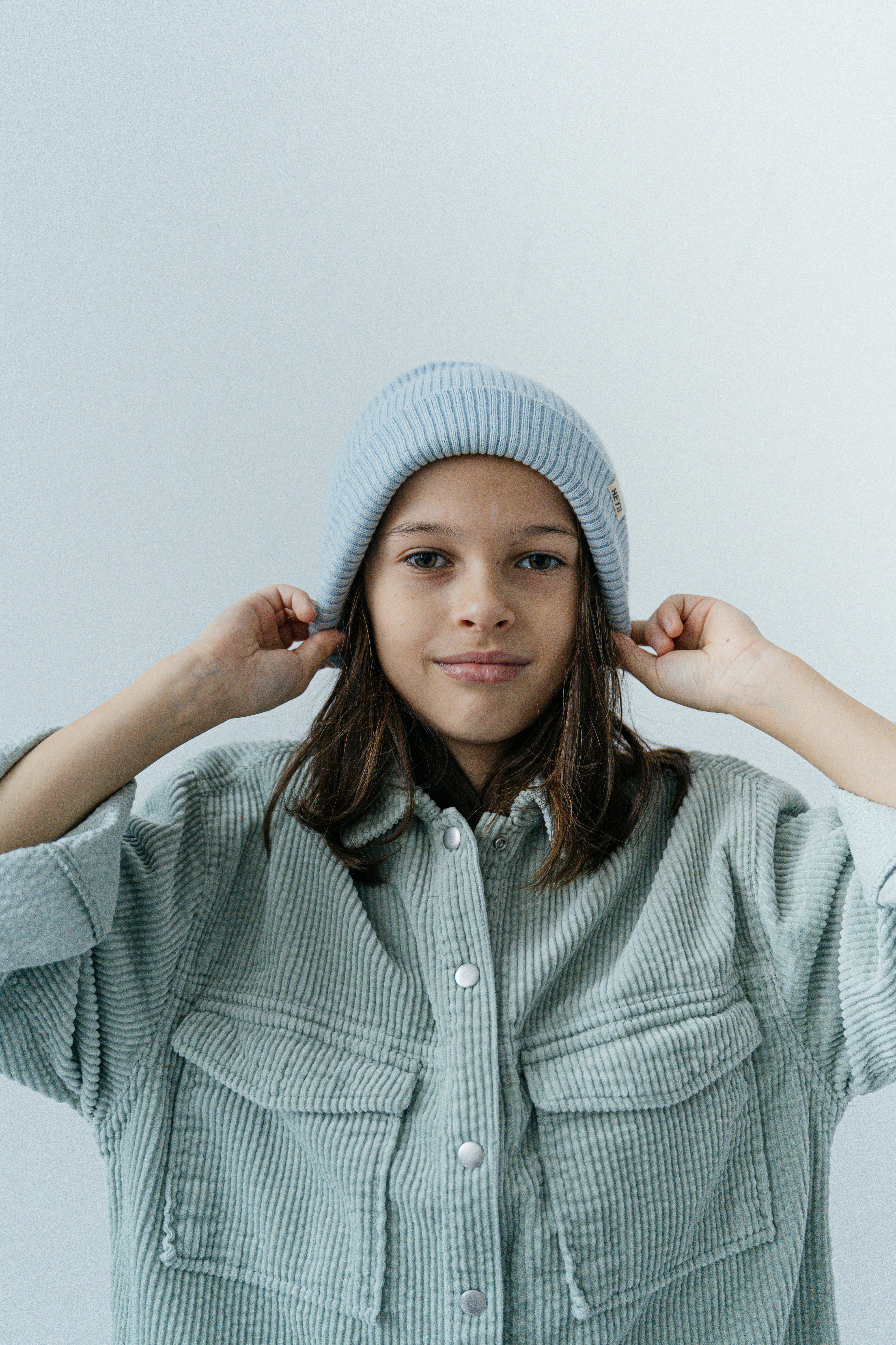 Charming portrait of a young girl wearing a stylish beanie and textured jacket in a neutral setting.