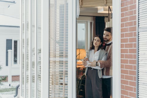 A couple stands warmly embracing near a large window inside a cozy home.
