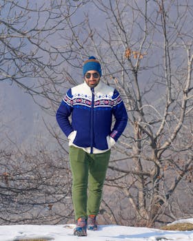 A stylish man in winter attire walking through a snowy, barren landscape with trees.
