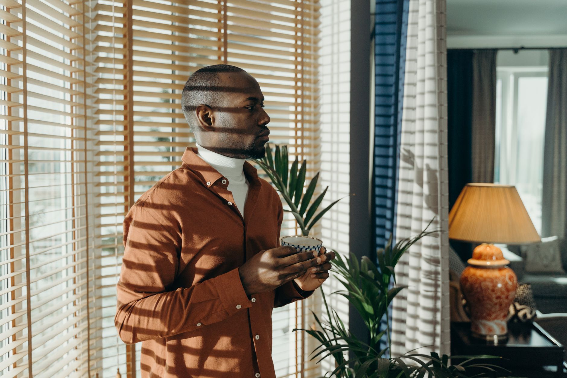 玻璃窗讓室內刺眼?直射光與漫射光一次搞懂 Man in brown shirt holding cup in sunlit room with blinds and plants.