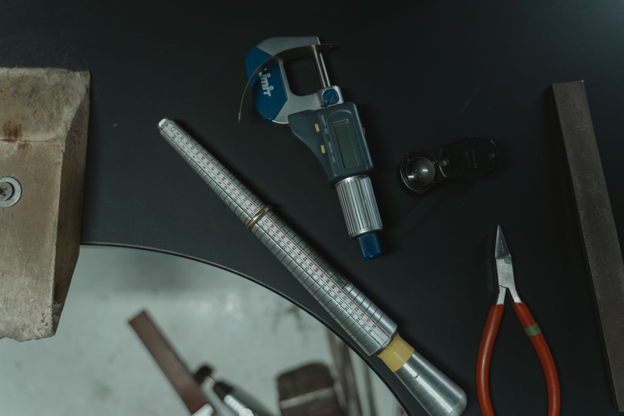 Jeweler's bench with a ring sizing mandrel holding a plain band, a digital micrometer, a loupe and pliers, used to measure carbon fiber and tungsten rings