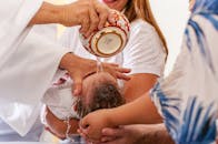 Crop anonymous priest pouring water on little baby head lying on parents hands during baptism ceremony in Catholic church