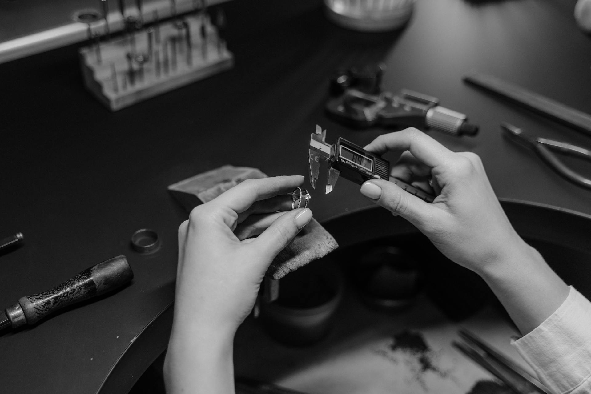 Black and white photo of a jeweler measuring a ring with a digital caliper in a workshop setting.