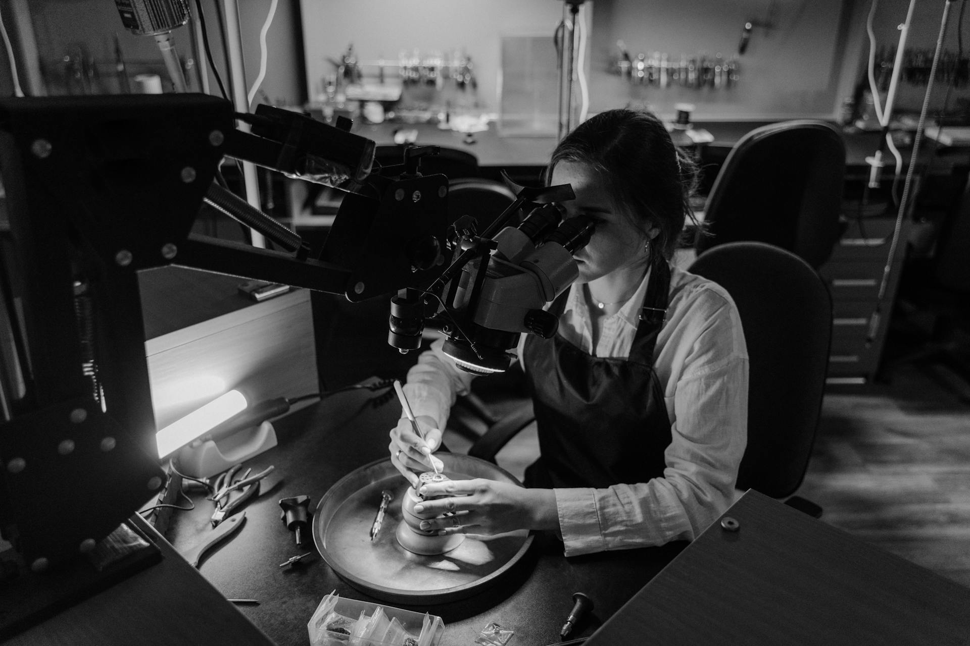 A skilled jeweler intensely focuses on crafting jewelry under a microscope in a workshop.