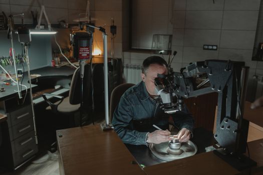 Jeweler focusing on intricate work using a microscope in a dimly lit workspace.
