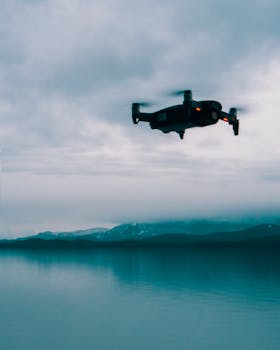A drone hovers over a tranquil sea with a distant coastline, under a cloudy sky.