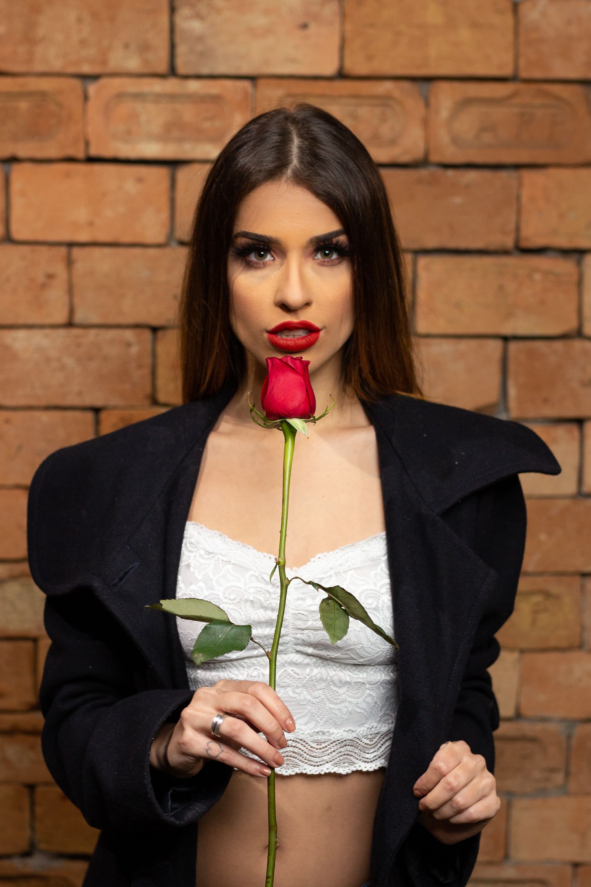 Beautiful woman in black coat with red rose posing against a rustic brick wall.