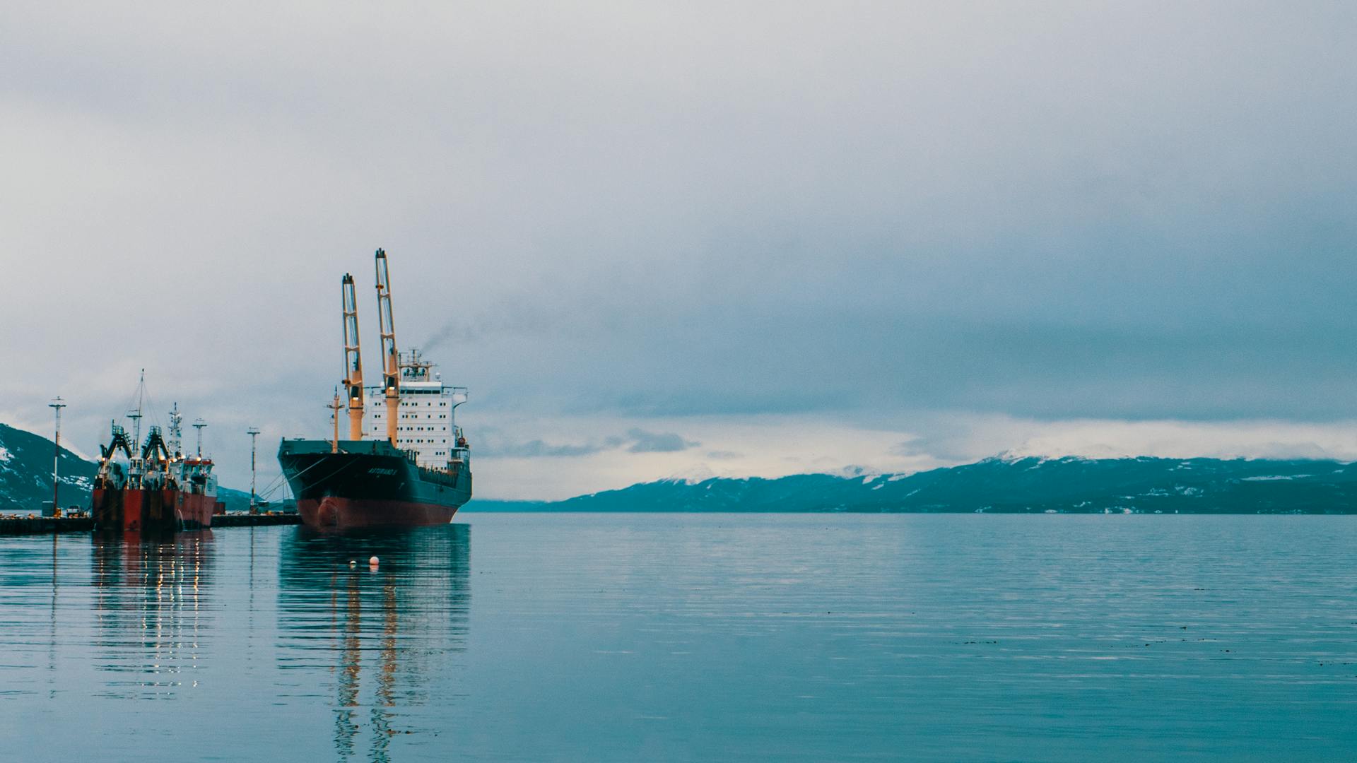Calm and serene harbor with cargo ships docked, surrounded by snowy mountains.