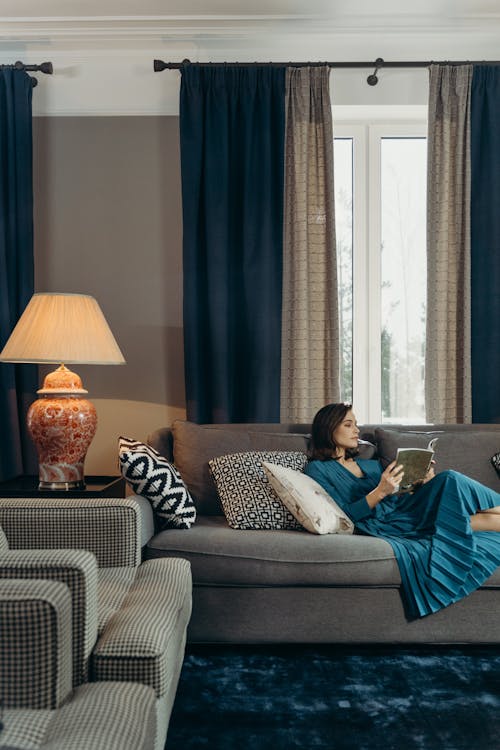 Woman in Blue Dress Sitting on Couch Near the Window