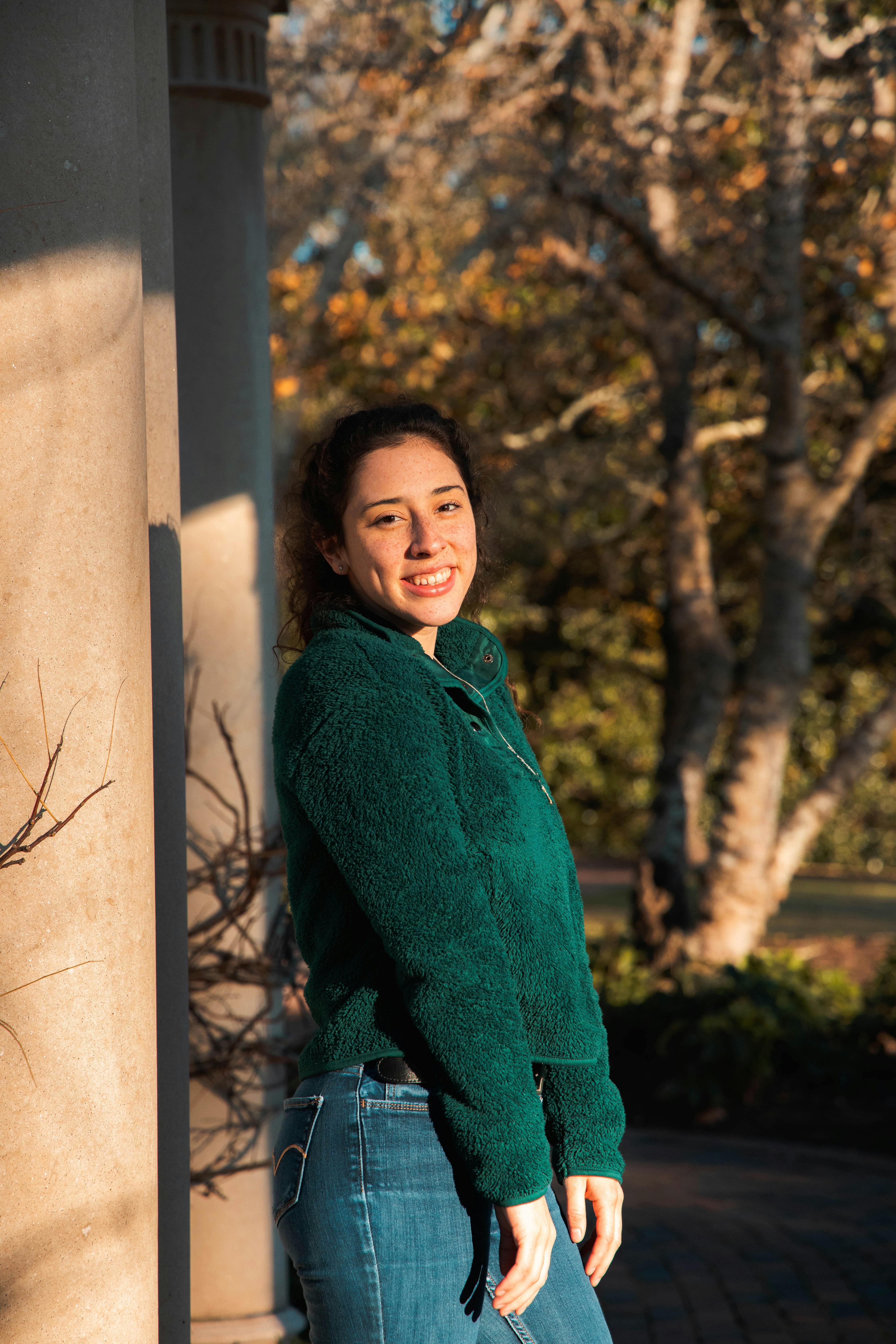 A cheerful woman leaning against a pillar, enjoying a sunny day outdoors in a green sweater.