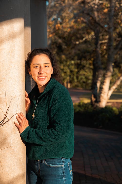 Woman in Green Sweater and Denim Jeans Smiling while Leaning on a Concrete Column