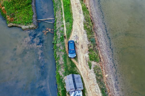 Drone view of automobile and near shabby house on narrow grassy ground surrounded with wet rice plantations in rural area