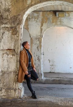 Stylish woman in a brown coat leans against a rustic archway, smiling outdoors.