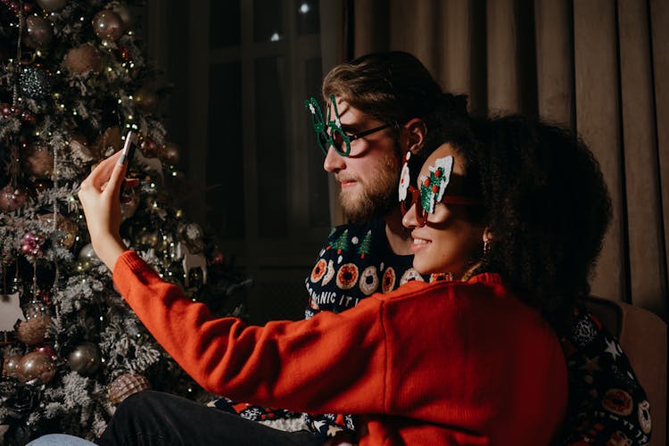 Side View Of A Man And A Woman Taking Picture Using A Smartphone Near A Christmas Tree