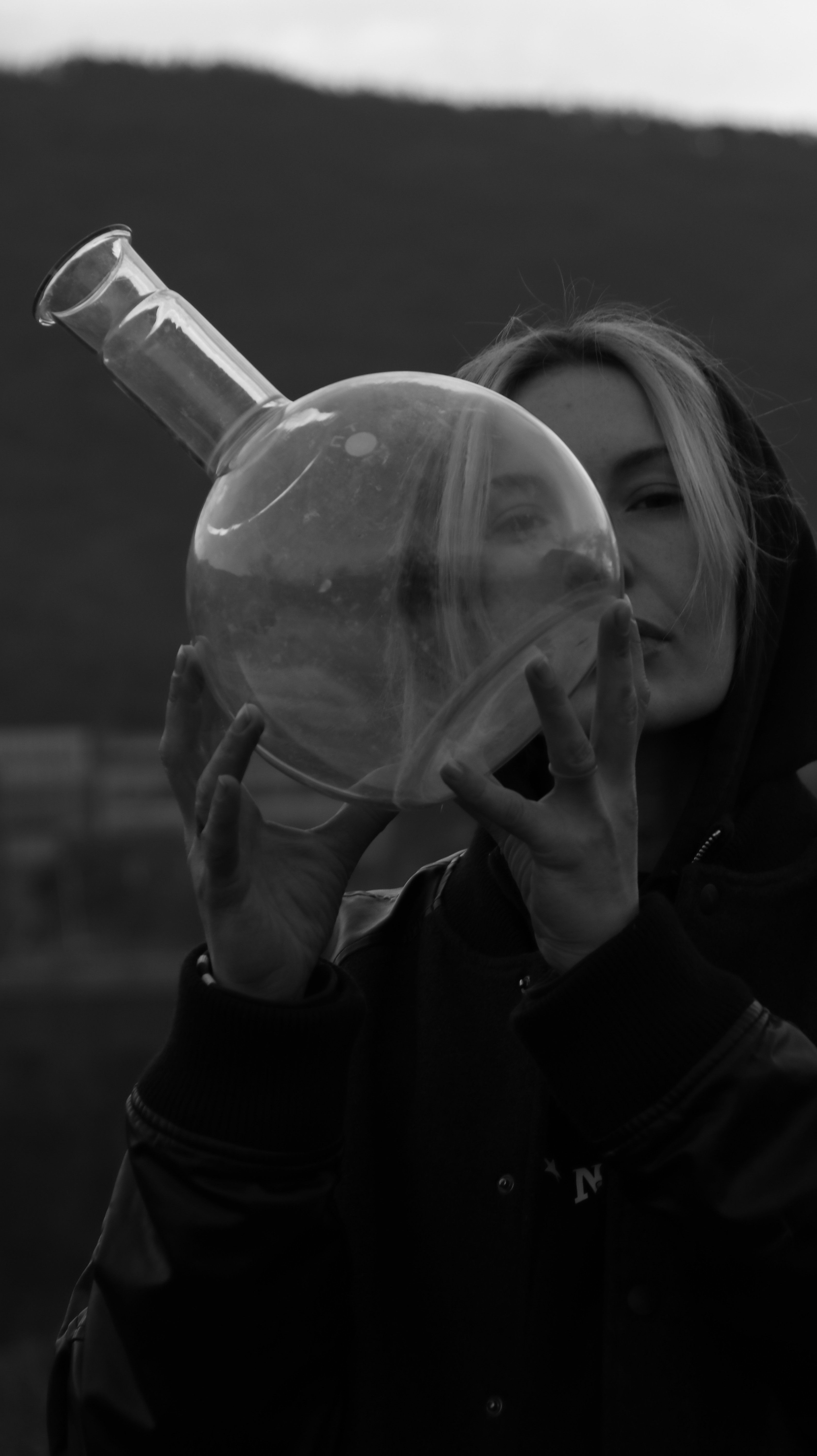 Woman in hoodie holding a clear glass flask in grayscale outdoors.