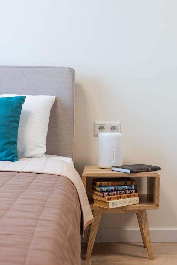 Stack of books and humidifier arranged on small minimalist wooden night table placed near comfortable bed with pillows in light room