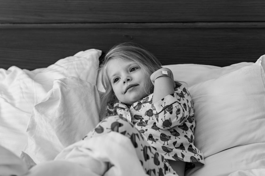 Free Black and white of adorable little girl in pajama lying on bed with soft pillows and looking away in sunny morning Stock Photo