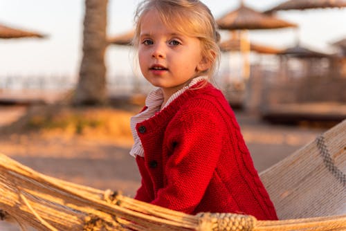 Free Side view crop cute little girl in stylish red cardigan sitting on hammock and looking at camera on sandy peaceful beach at sunset Stock Photo