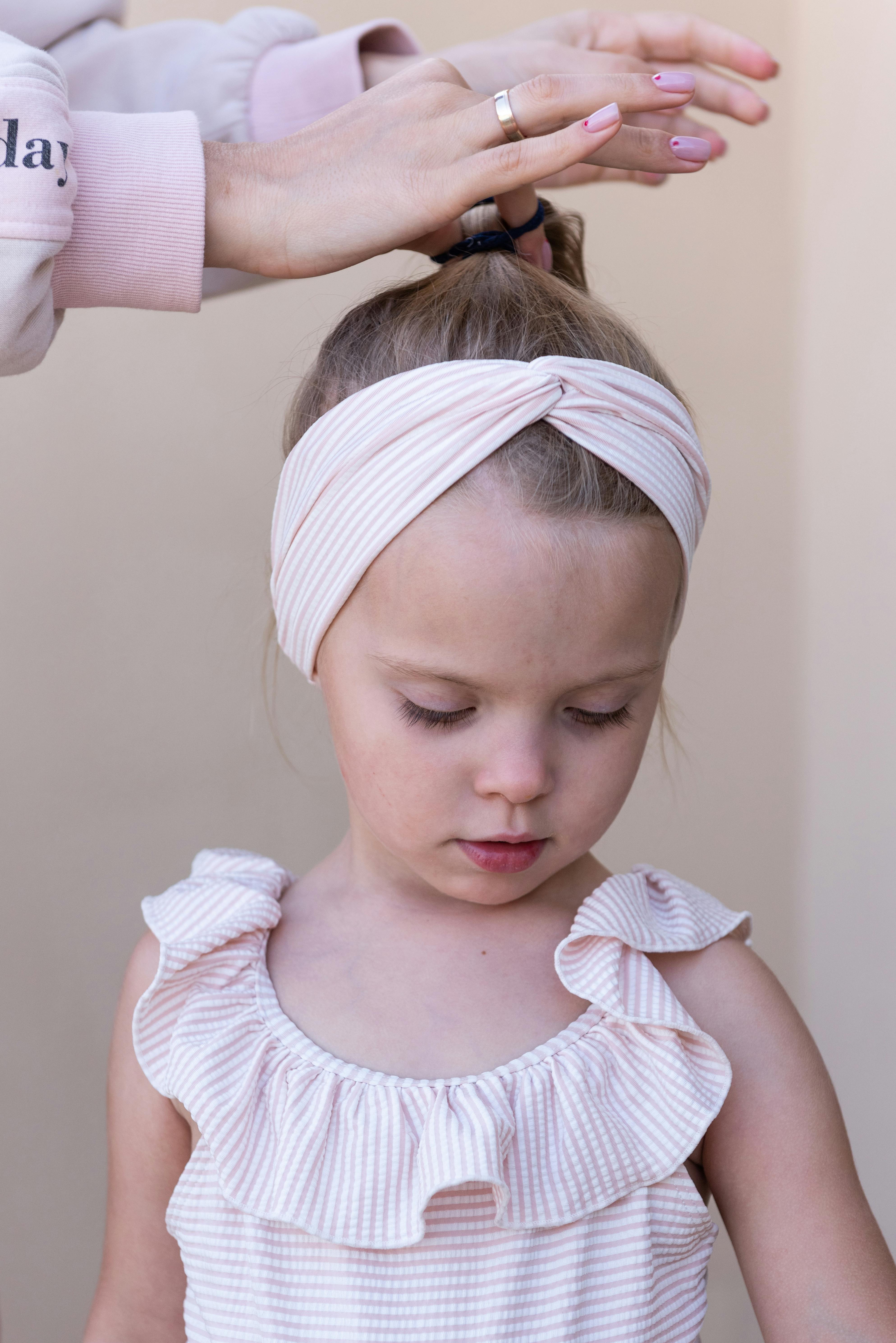 Caring mother doing ponytail hairstyle for adorable little daughter wearing stylish sundress and headband in light room