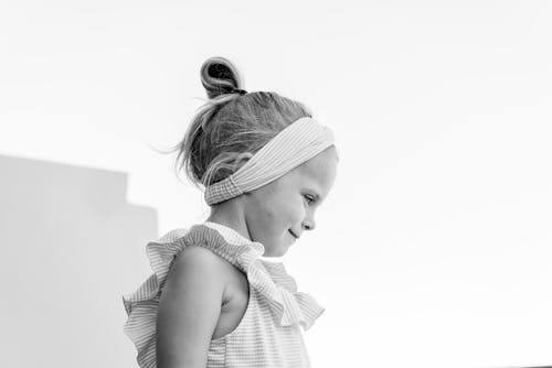 Black and white adorable content little girl wearing stylish sundress and headband standing under clear sky and looking away dreamily