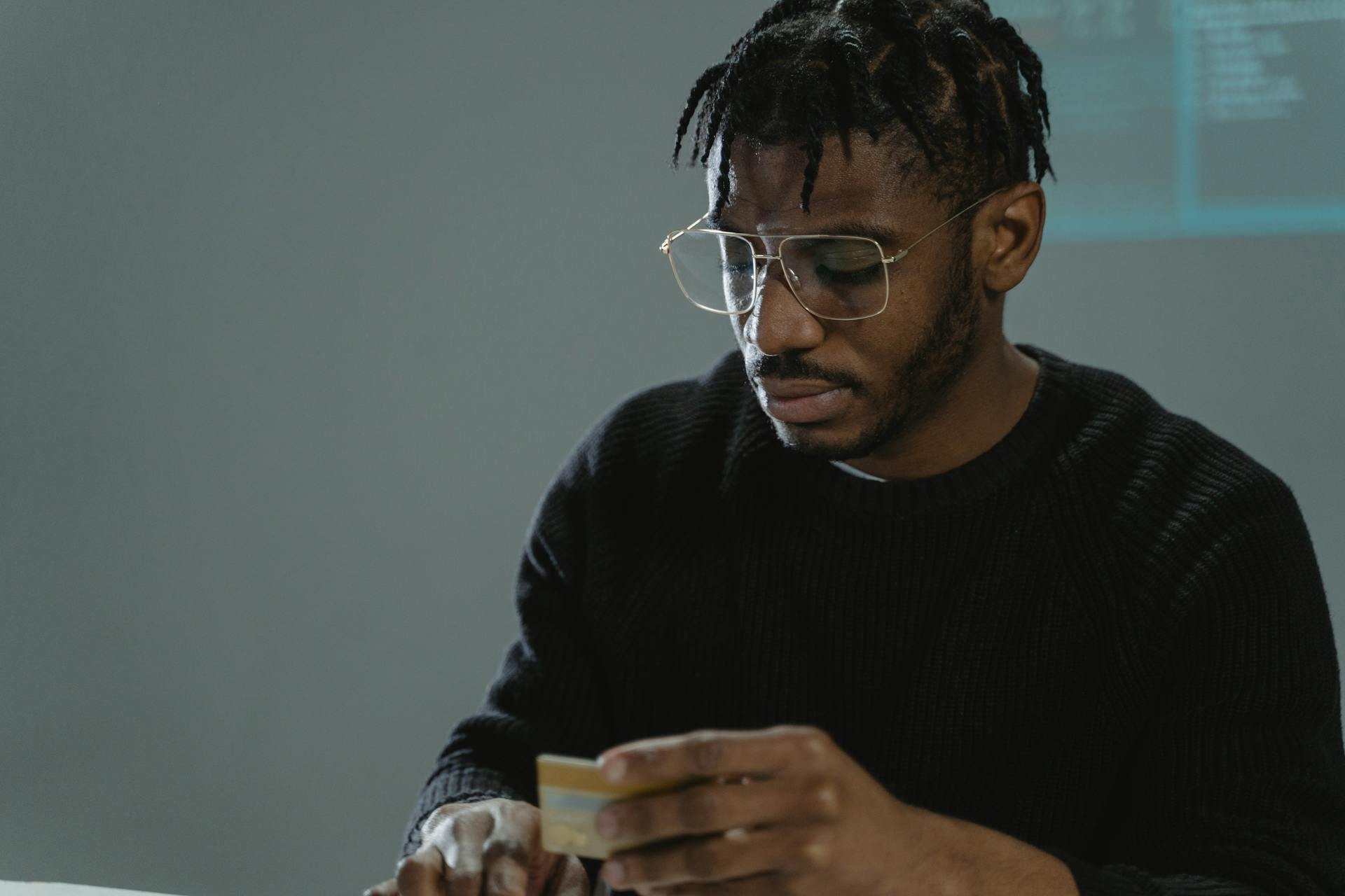 Focused man with eyeglasses holding a credit card in a dimly lit room, wearing a black sweater.