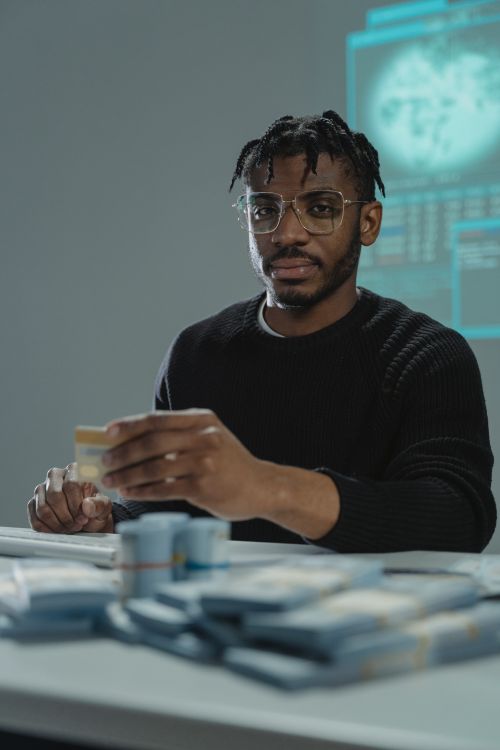 Free A seated man in glasses and sweater holding a credit card near stacks of cash, suggesting financial themes. Stock Photo