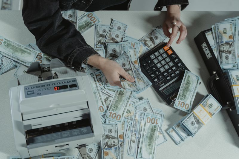 Hands counting 100 dollar bills using a calculator and money counter on a table.