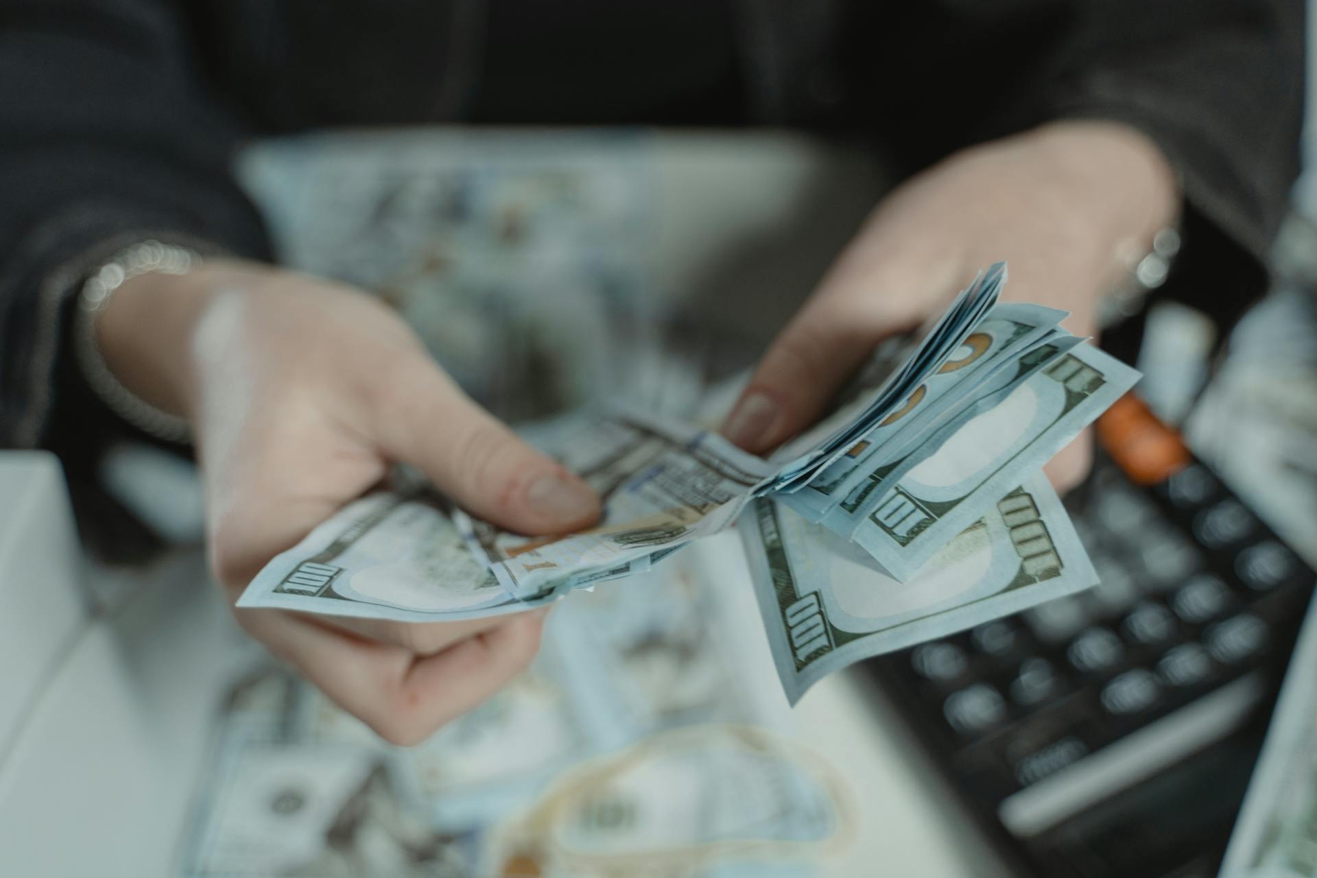 Close-up of hands counting hundred dollar bills with a calculator in the background.