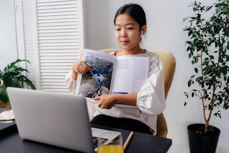 Girl Holding Book In Front Of Silver Laptop