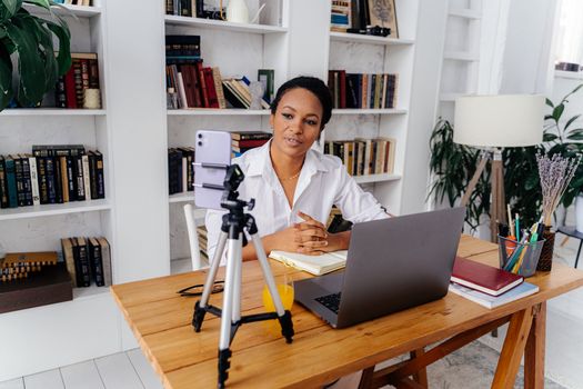 A woman working remotely using a laptop and smartphone in a home office setup with bookshelves.