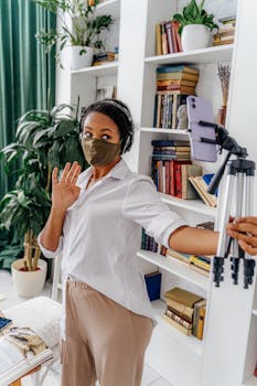 A woman with a mask uses a smartphone on a tripod for a recorded virtual communication.