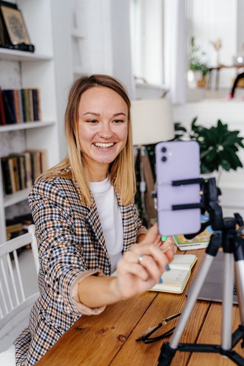 Free Smiling woman in a contemporary home office using a smartphone on a tripod for a video call. Stock Photo