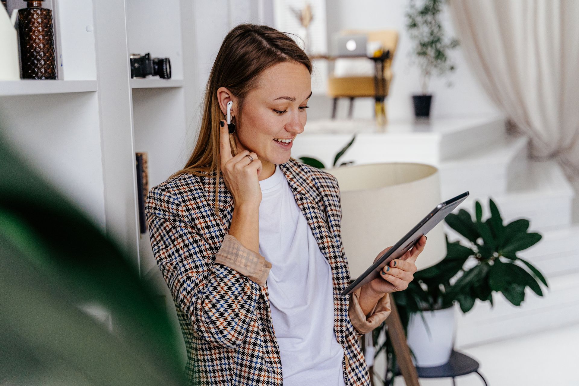 Smiling woman with earbuds interacting with a tablet in a cozy, modern room with plants.