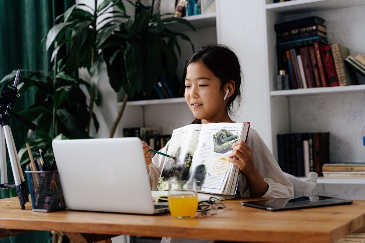 A school-age student studying at home with a book and laptop during an online lesson.