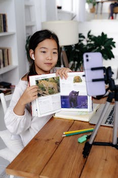 Young girl displays book during online class using smartphone and tripod.