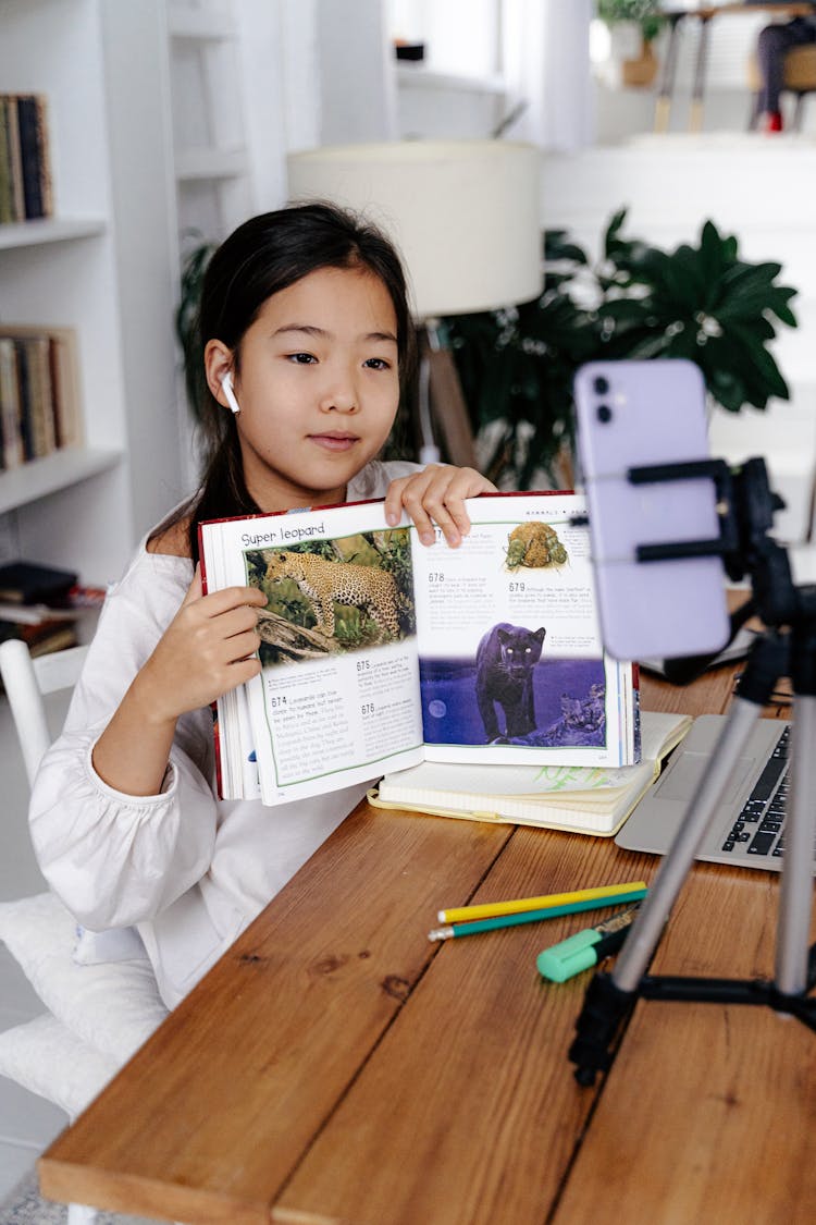 Young Girl Recording Herself With A Phone On A Tripod And Showing A Book To The Camera