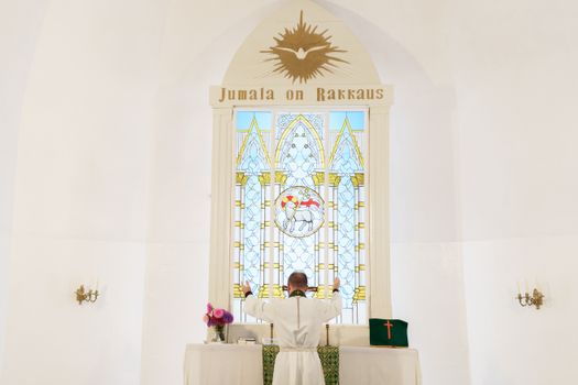 A pastor stands at an altar, leading a prayer in a beautifully lit church interior with stained glass windows.